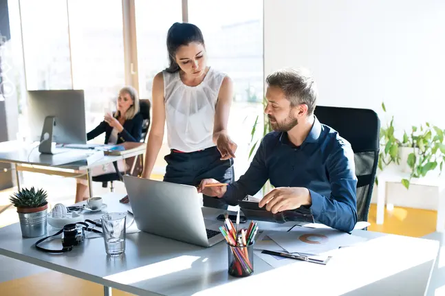 A man at a desk works on a laptop while a woman stands next to him